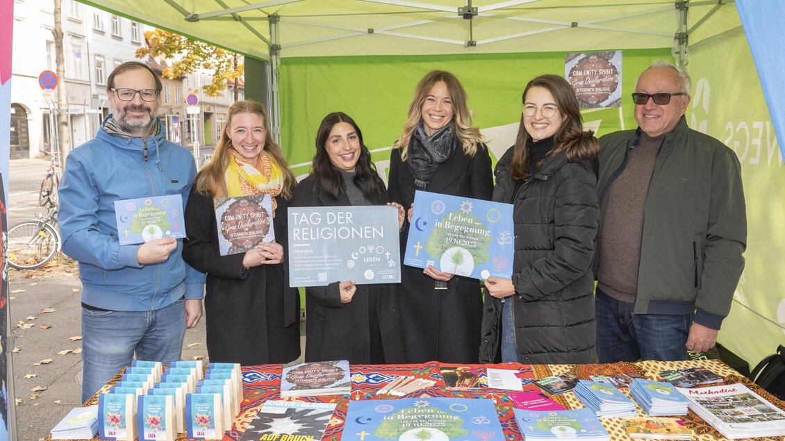 Sechs Mitglieder des Organisationsteams stehen an einem Infostand und präsentieren Plakate zum Tag der Religionen in Graz. | © Fotos Fischer
