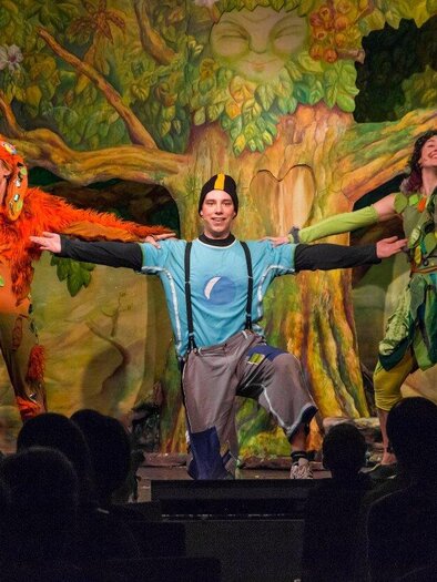 Drei fröhliche Schauspieler in bunten Kostümen posieren vor einer lebhaften Kulisse. Im Hintergrund sind Gebäude mit orientalischen Türmen und ein märchenhafter Weg zu sehen. | © Theater Heuschreck
