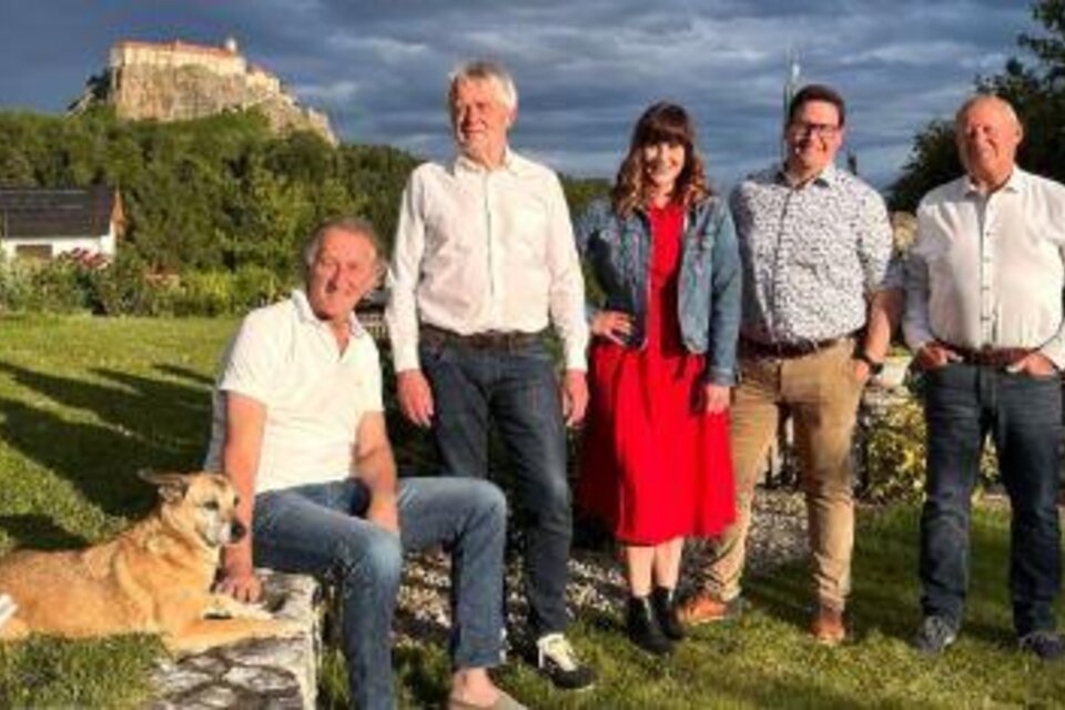 Eine Gruppe von fünf Personen steht in einem grünen Garten. Im Vordergrund sitzt ein Hund, während im Hintergrund eine Burg sichtbar ist. | © © Die Hausband