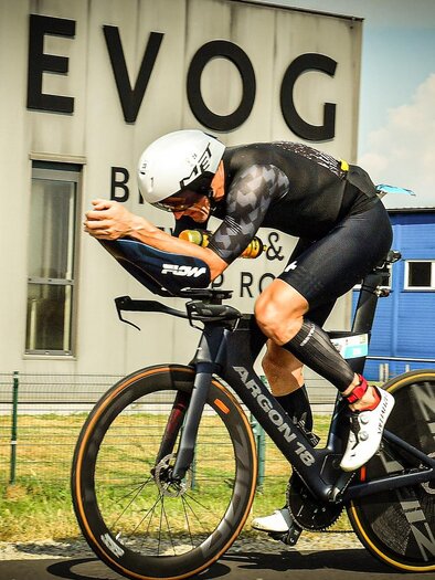 A cyclist in racing gear is riding on a road in front of a building. In the background, the "BEVOG" logo is visible. | © team triaguide