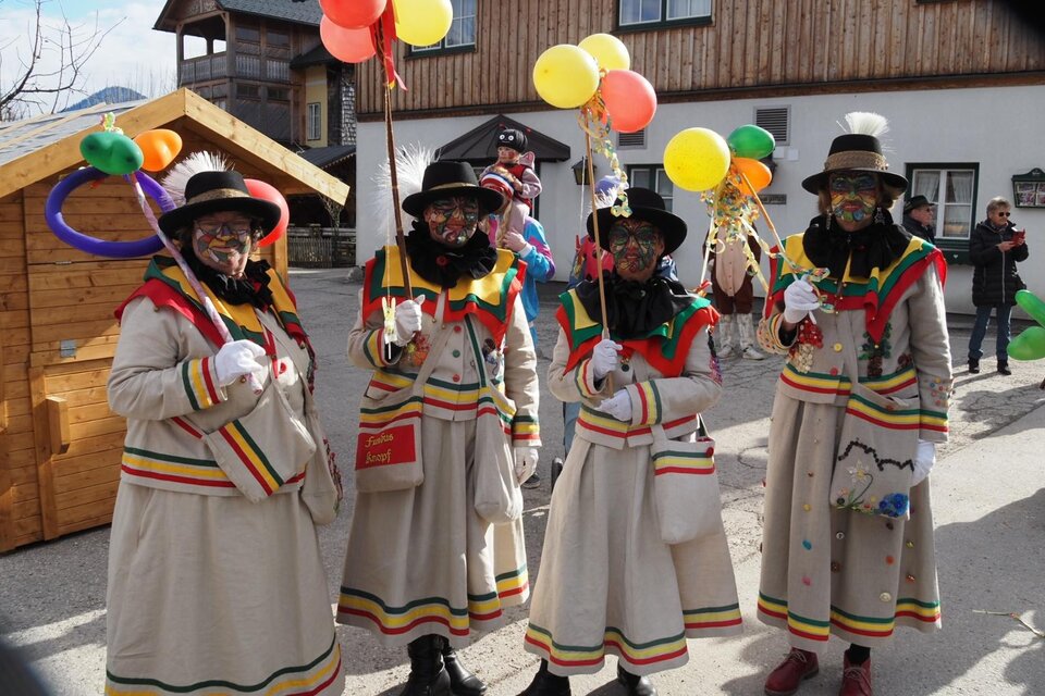 Vier Knopferl in ihrem traditionellen Gewand stehen in einer festlichen Faschingsatmosphäre im Herzen von Altaussee und halten bunte Luftballons. Im Hintergrund sind Holzbauten und weitere Feiernde zu sehen. | © Theresa Schwaiger