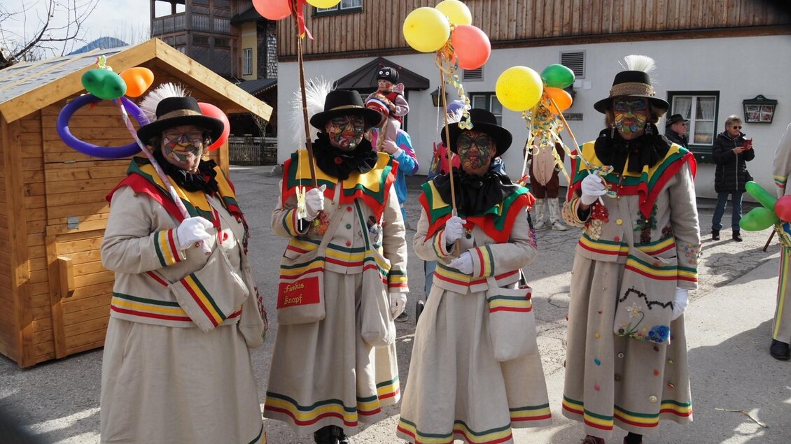 Vier Knopferl in ihrem traditionellen Gewand stehen in einer festlichen Faschingsatmosphäre im Herzen von Altaussee und halten bunte Luftballons. Im Hintergrund sind Holzbauten und weitere Feiernde zu sehen. | © Theresa Schwaiger
