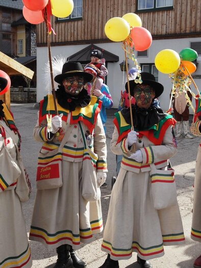 Four little "Knopferl", traditional carnival figures in the Ausseerland Salzkammergut region, in their characteristicl carnival attire. They stand in a festiv atmosphere in the heart of Altaussee, holding colorful balloons. In the background, wooden structures and more partygoers can be seen. | © Theresa Schwaiger