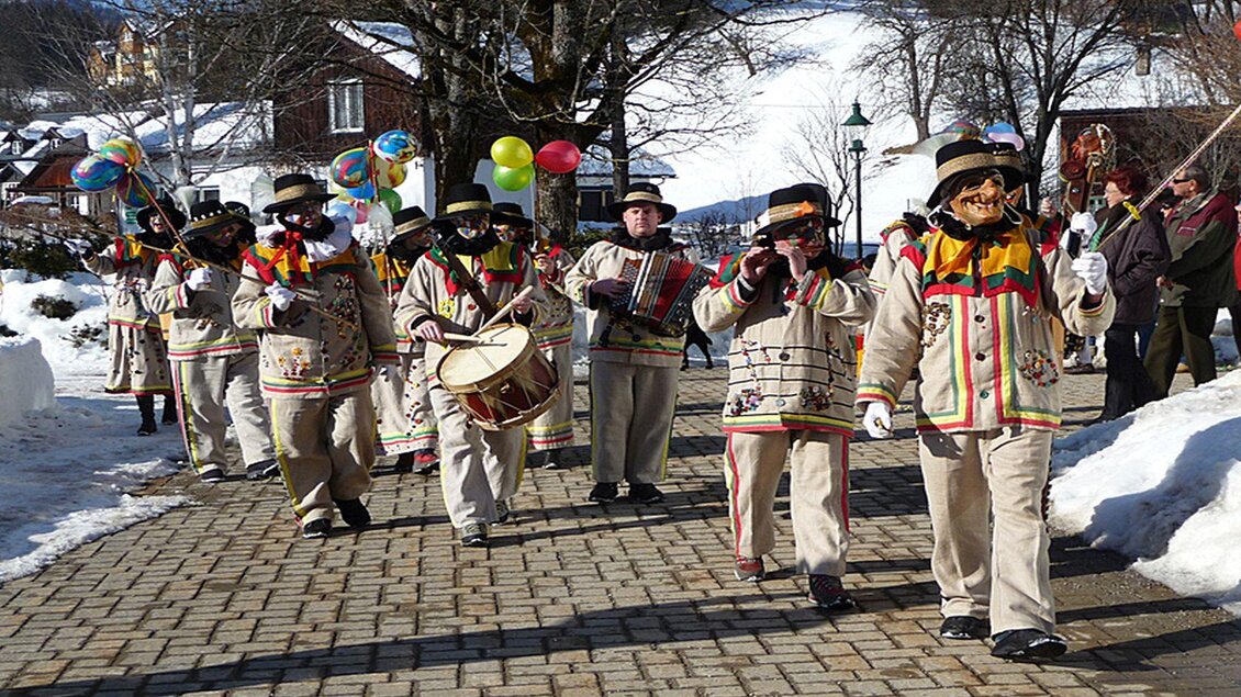 Eine fröhliche Gruppe von Altausseer Knopferln in ihren traditionellen, symbolträchtigen Gewändern, die zum Ausseer Fasching an ihrem Paradiestag durch eine schneebedeckte Straße marschiert. Sie schwingen ihre Angeln, spielen Trommeln und Instrumente und tragen bunte Girlanden und Luftballons. | © Hermann Schröttenhamer
