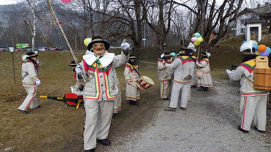 Eine Gruppe von Altausseer Knopferln in ihren traditionellen Gewändern, die zum Ausseer Fasching an ihrem Paradiestag durch Altaussee marschieren und Musik und Freude verbreiten. Dabei schwingen sie ihre Angeln, schlagen Trommeln, spielen Instrumente und tragen bunte Girlanden und Luftballons herum. | © Hermann Schröttenhamer