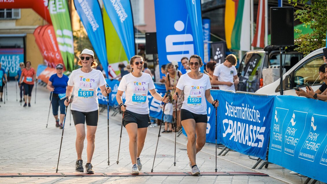 Drei Frauen gehen mit Nordic-Walking-Stöcken bei einer Veranstaltung. Im Hintergrund sind bunte Banner und Teilnehmer zu sehen. | © Hikimus