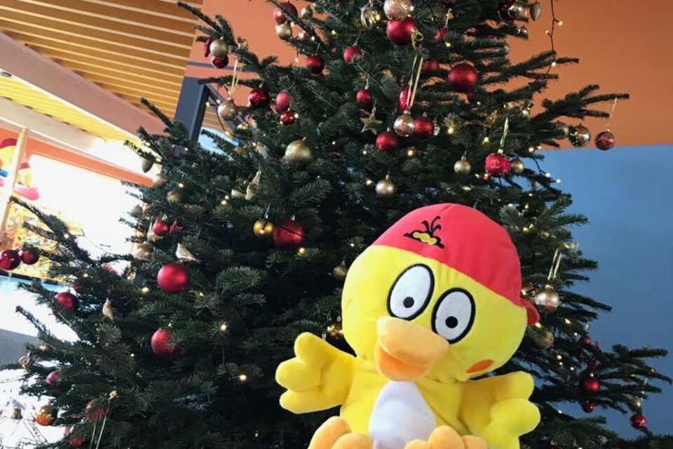 Ein festlich geschmückter Weihnachtsbaum mit funkelnden Lichtern und roten sowie goldenen Kugeln sorgt für stimmungsvolles Ambiente. Davor sitzt ein fröhliches gelbes Maskottchen auf einem weihnachtlich verpackten Geschenk und verbreitet gute Laune in der H2O-Therme in Bad Waltersdorf. | © H2O-Hoteltherme GmbH