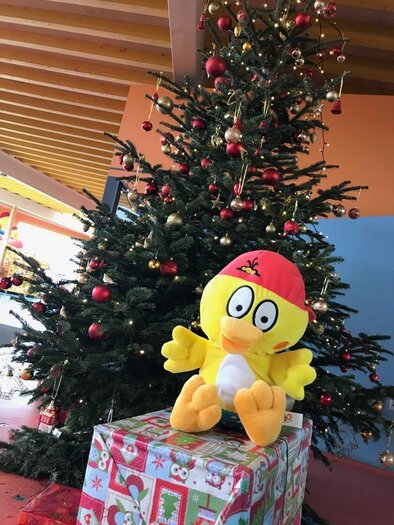A festively decorated Christmas tree with sparkling lights and red as well as golden ornaments creates a cheerful atmosphere. In front of it sits a cheerful yellow mascot on a Christmas-wrapped gift, spreading good cheer in the H2O Therme in Bad Waltersdorf. | © H2O-Hoteltherme GmbH