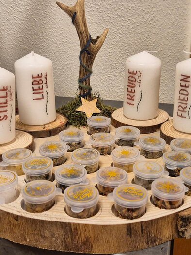 Eine dekorative Holzplatte mit mehreren Kerzen und kleinen Behältern. Die Kerzen tragen die Wörter "Stille," "Liebe," "Freude," und "Frieden." | © Marion Gallautz