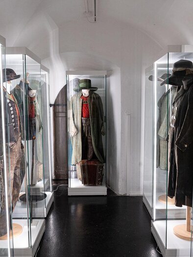 Eine Frau steht im Vordergrund und breitet die Arme aus, während vier Männer hinter ihr stehen. Der Hintergrund ist dunkel mit grafischen Mustern. | © Stadtmuseum Trofaiach