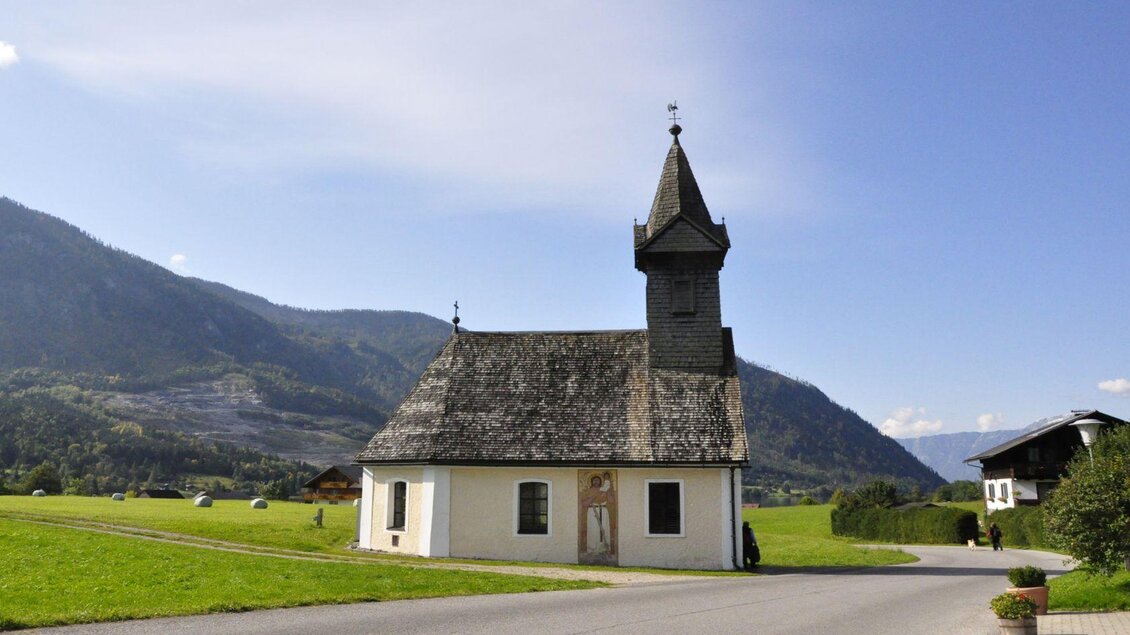 Eine charmante Kapelle mit einem spitzen Turm, umgeben von grünen Wiesen und Bergen im Hintergrund. Der Himmel ist klar und blau. | © Steinegger