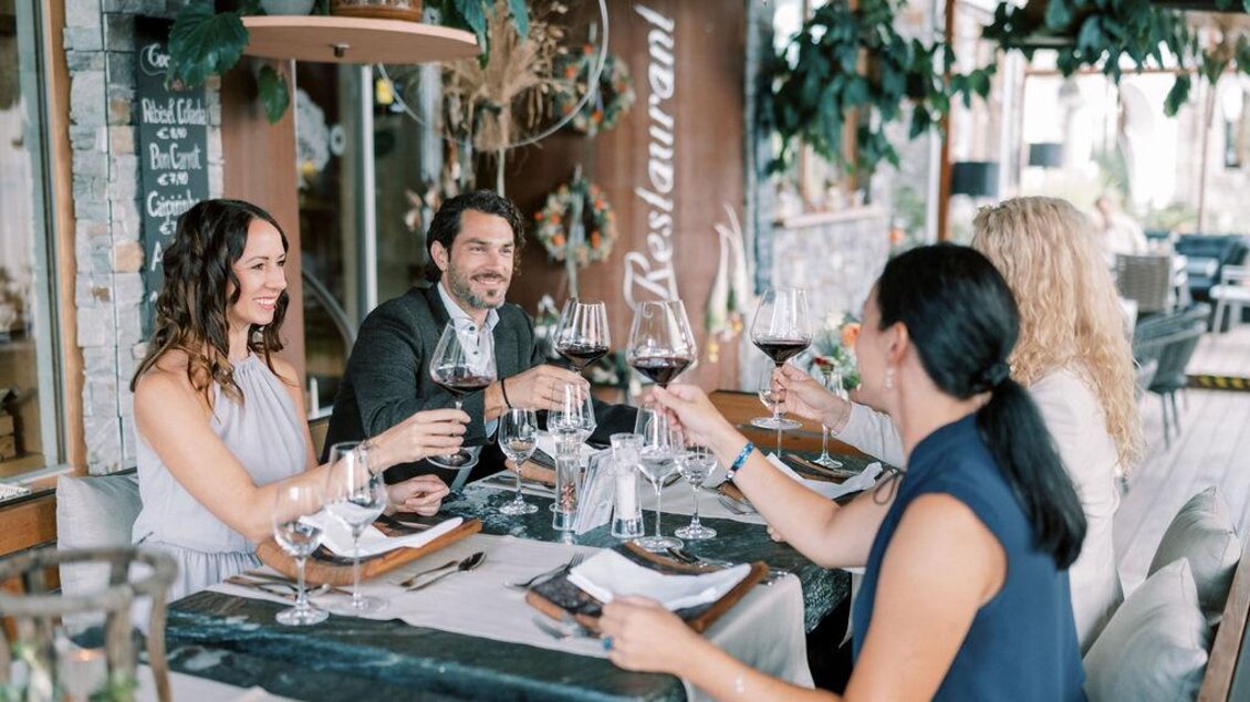Eine Gruppe von Freunden sitzt an einem Tisch in einem Restaurant und stößt mit Weingläsern an. Die Atmosphäre ist gemütlich und einladend, umgeben von Pflanzen und einem modernen Ambiente. | © Garten Hotel Ochensberger