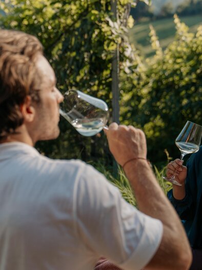 A gentleman and a lady enjoy a glass of white wine in the vineyard between the rows of grapevines in the sunshine at Kehlberg, Graz. | © studio draussen