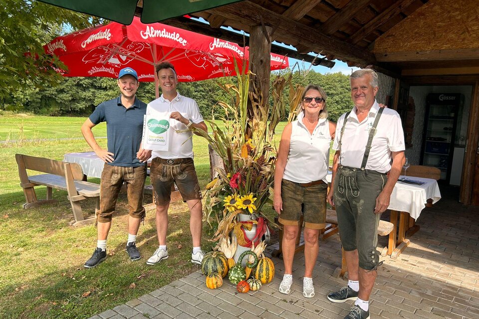 Eine Gruppe von vier Personen steht lächelnd vor einem rustikalen Gartenrestaurant. Im Hintergrund sind bunte Dekorationen und ein Sonnenschirm zu sehen. | © Golf Resort Klöch