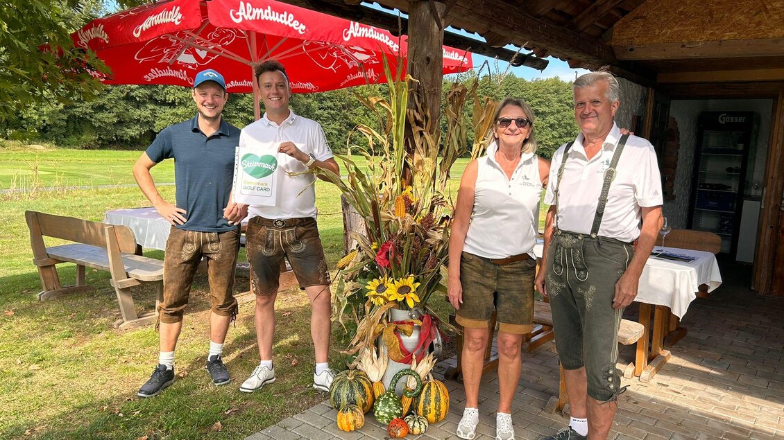 Eine Gruppe von vier Personen steht lächelnd vor einem rustikalen Gartenrestaurant. Im Hintergrund sind bunte Dekorationen und ein Sonnenschirm zu sehen. | © Golf Resort Klöch