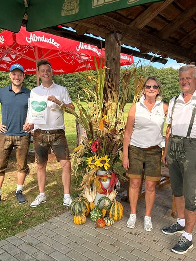 Eine Gruppe von vier Personen steht lächelnd vor einem rustikalen Gartenrestaurant. Im Hintergrund sind bunte Dekorationen und ein Sonnenschirm zu sehen. | © Golf Resort Klöch