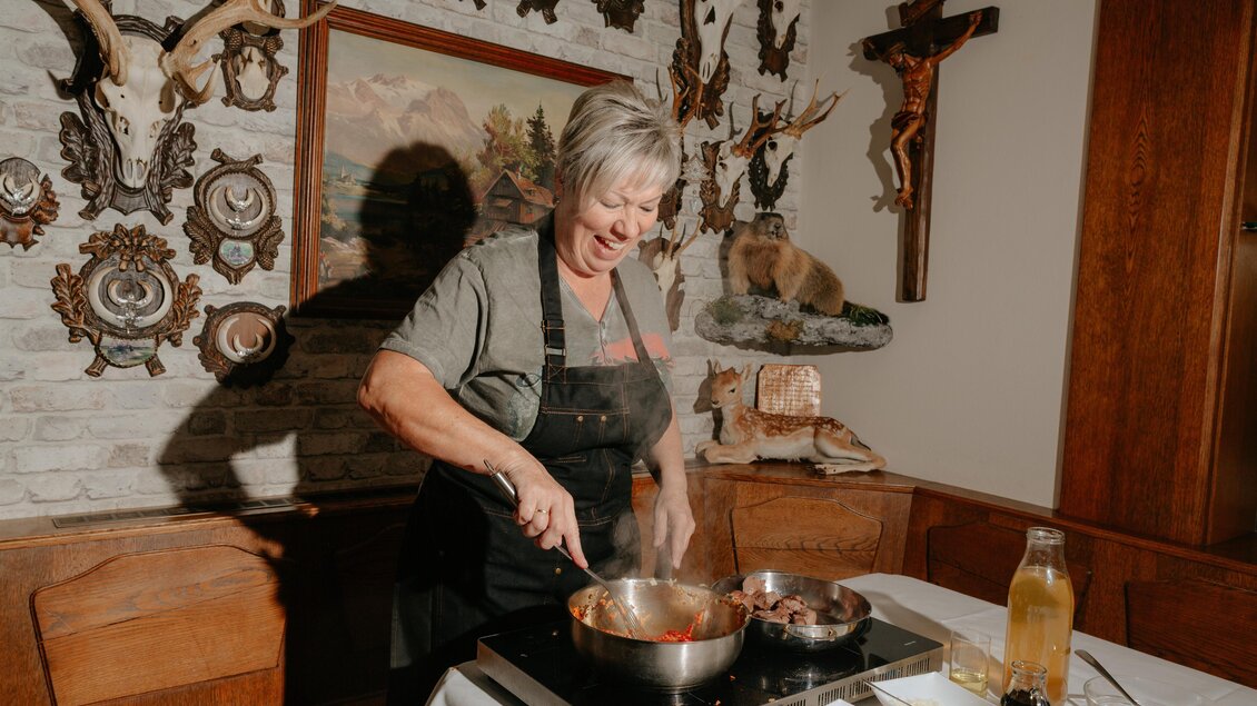 Eine lachende Frau kocht in einer gemütlichen Umgebung. Im Hintergrund sind dekorative Elemente und antike Bilder zu sehen. | © Studio-draussen