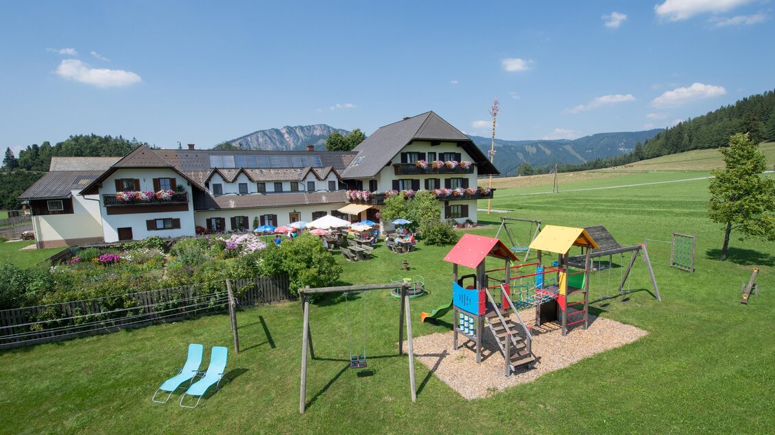 Ein malerisches Ferienhaus mit einem Spielplatz im Vordergrund. Im Hintergrund erstrecken sich grüne Wiesen und Berge unter einem klaren blauen Himmel. | © GH Frankenhof