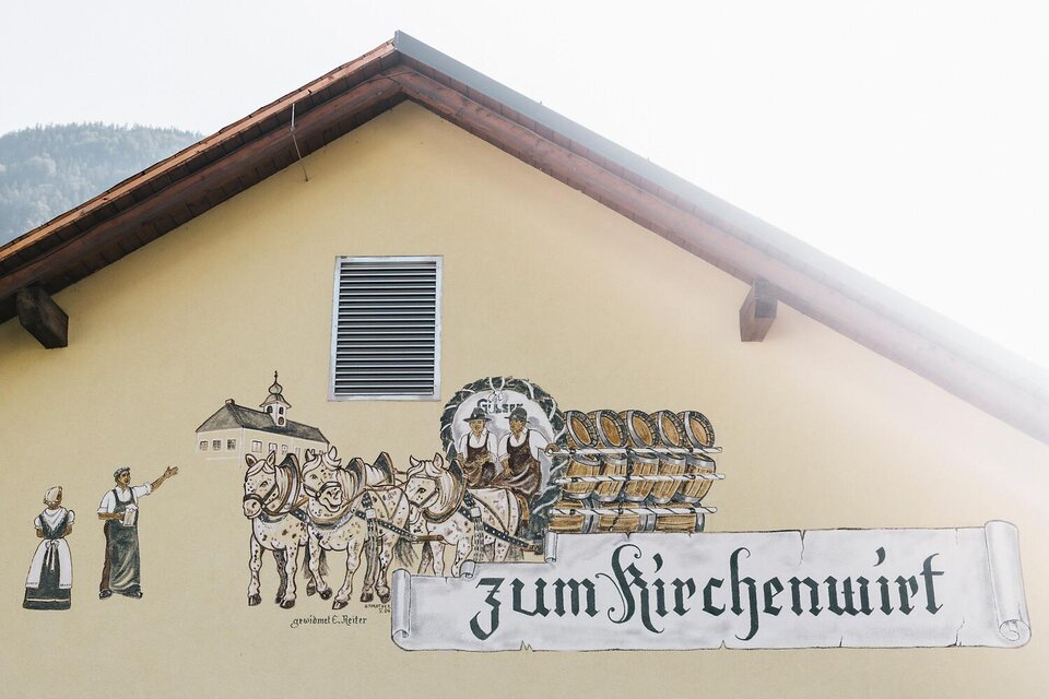 Eine Wandmalerei an einem Gebäude zeigt Menschen in traditioneller Kleidung und ein von Pferden gezogenes Gefährt. Darüber steht der Schriftzug "zum Kirchenwirt". | © TV Gesäuse