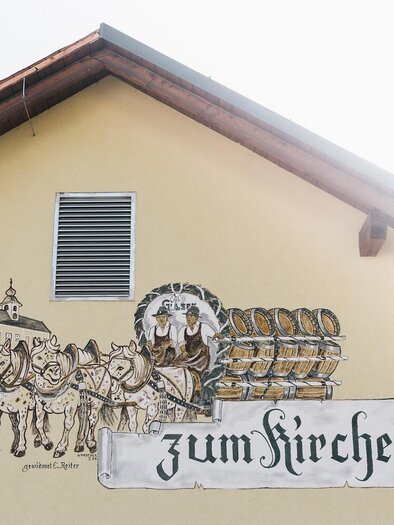 Eine Wandmalerei an einem Gebäude zeigt Menschen in traditioneller Kleidung und ein von Pferden gezogenes Gefährt. Darüber steht der Schriftzug "zum Kirchenwirt". | © TV Gesäuse