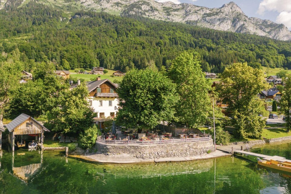 Der malerischer Grundlsee mit klar, grünem Wasser umgeben von Bäumen und den Bergen des Ausseerlandes. Im Vordergrund steht das gemütliche Dorfwirtshaus Stöckl. | © Karl Steinegger