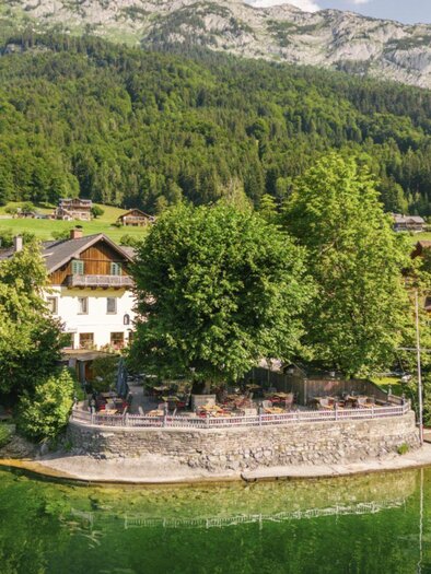 Der malerischer Grundlsee mit klar, grünem Wasser umgeben von Bäumen und den Bergen des Ausseerlandes. Im Vordergrund steht das gemütliche Dorfwirtshaus Stöckl. | © Karl Steinegger