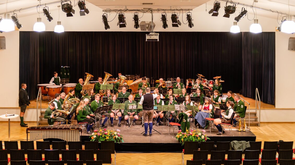 Eine Musikkapelle probt auf einer Bühne in einem großen Saal. Im Hintergrund sind zahlreiche Musiker in Uniform und verschiedene Instrumente zu sehen. | © David Stocker