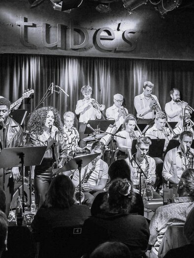 A large band with several musicians is playing on a stage. The audience is sitting in front and enjoying the concert in a cozy atmosphere. | © Johannes Teichert
