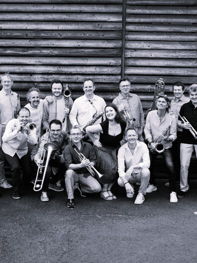 A large group of musicians stands in front of a wooden wall. They hold various musical instruments, including trumpets and trombones. | © Big Band Bad Gleichenberg