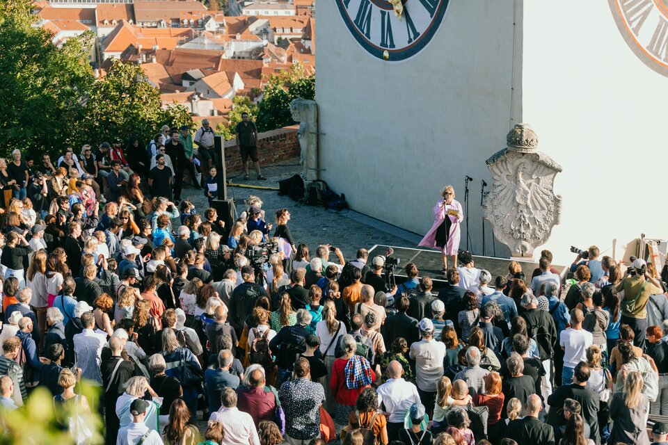 Eine große Menschenmenge versammelt sich vor einer Bühne mit einer Uhr im Hintergrund. Eine Person steht auf der Bühne und spricht zu den Anwesenden. | © steirischer herbst-Johanna Lamprecht