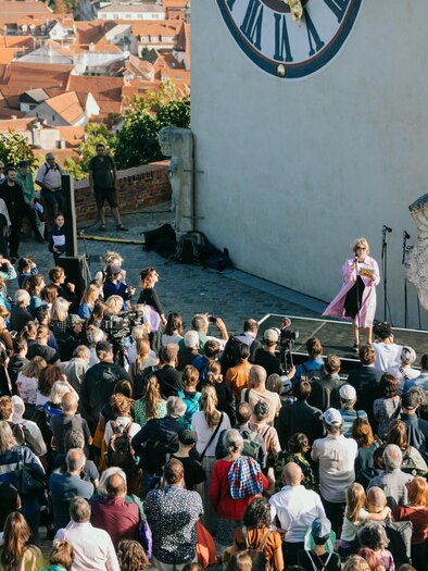 A large crowd gathers in front of a stage with a clock in the background. A person stands on the stage and speaks to the attendees. | © steirischer herbst-Johanna Lamprecht