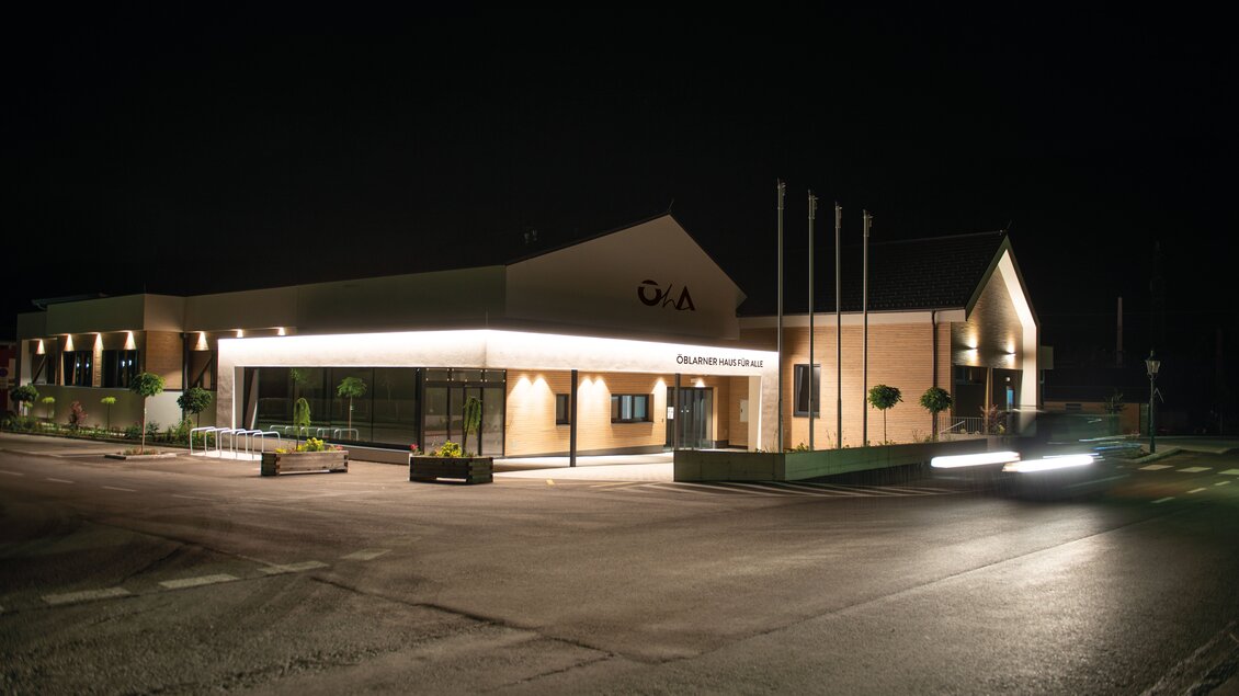 Ein modernes Gebäude mit beleuchtetem Eingangsbereich in der Nacht. Es sind einige Pflanzen und Flaggen im Vordergrund zu sehen. | © Marktgemeinde Öblarn