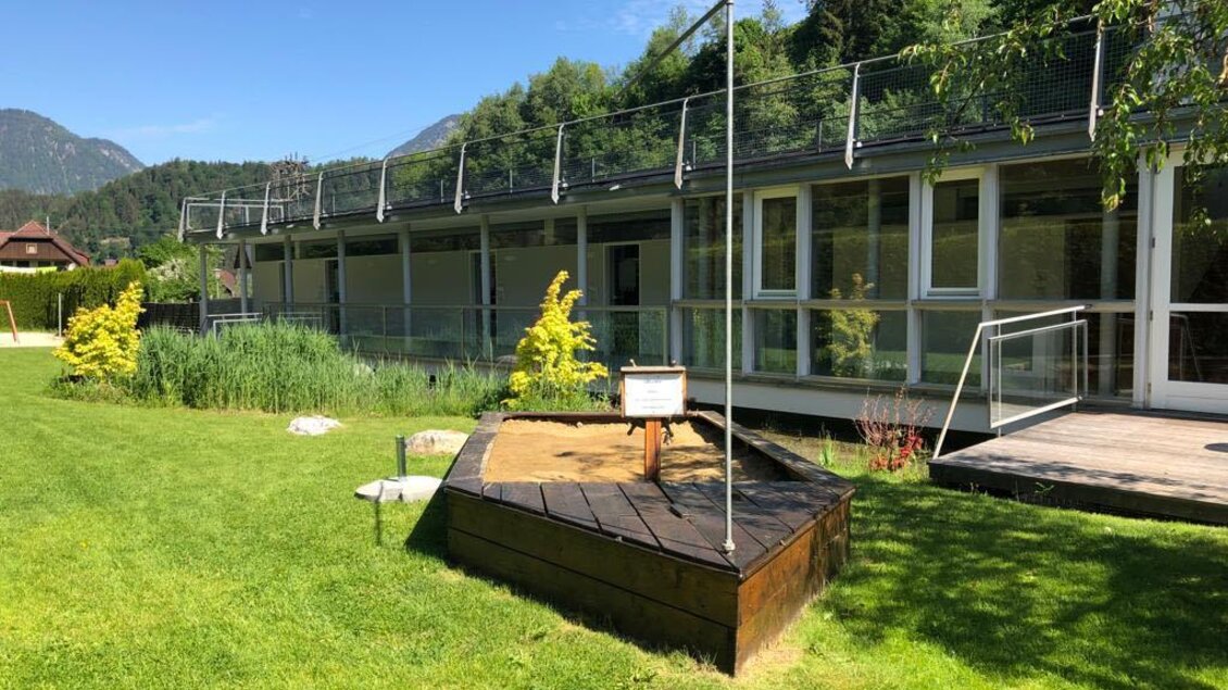 Ein moderner Gebäude mit großen Fenstern, umgeben von Grünflächen. Im Vordergrund befindet sich ein Sandkasten. | © Schwimmbadbuffet 