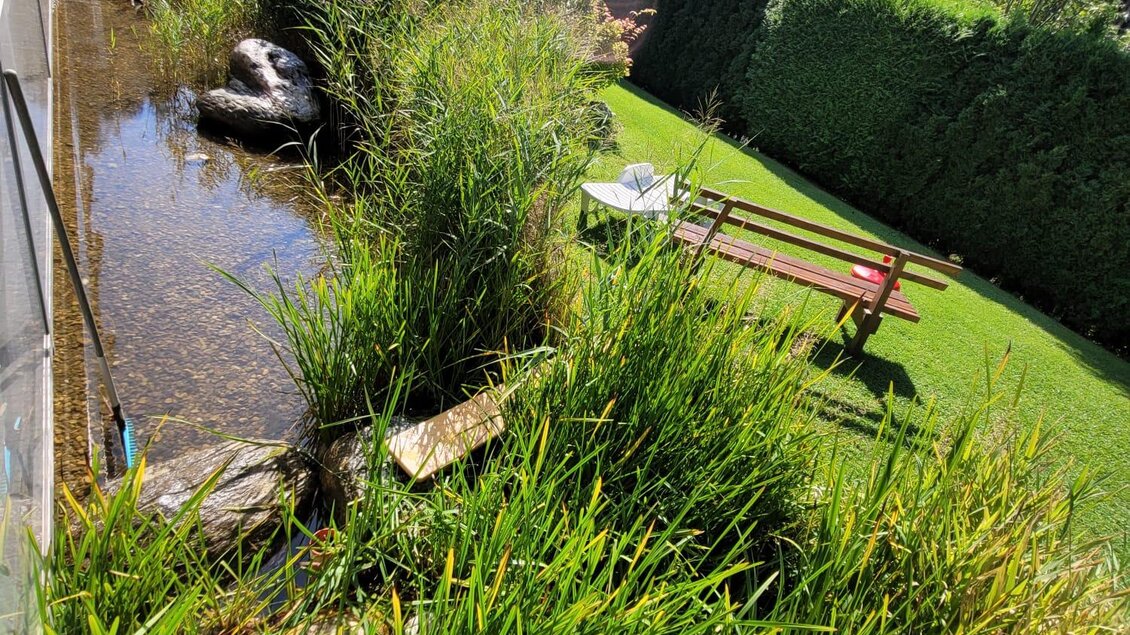 Ein ruhiger Garten mit hohem Gras, einem kleinen Gewässer und einer Parkbank. Im Hintergrund sind sanfte Hügel und Bäume sichtbar. | © Tim Fuchsberger 