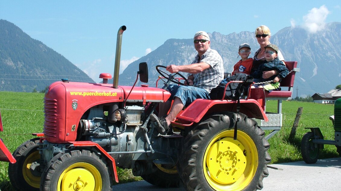 Ein roter Traktor fährt auf einer Landstraße mit einer Familie darauf. Im Hintergrund sind Berge und ein blauer Himmel zu sehen. | © Pürcherhof