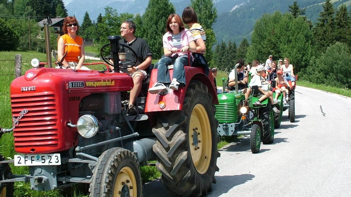 Ein roter Traktor mit mehreren Personen fährt auf einer ländlichen Straße. Im Hintergrund sind grüne Wälder und Berge zu sehen. | © Pürcherhof