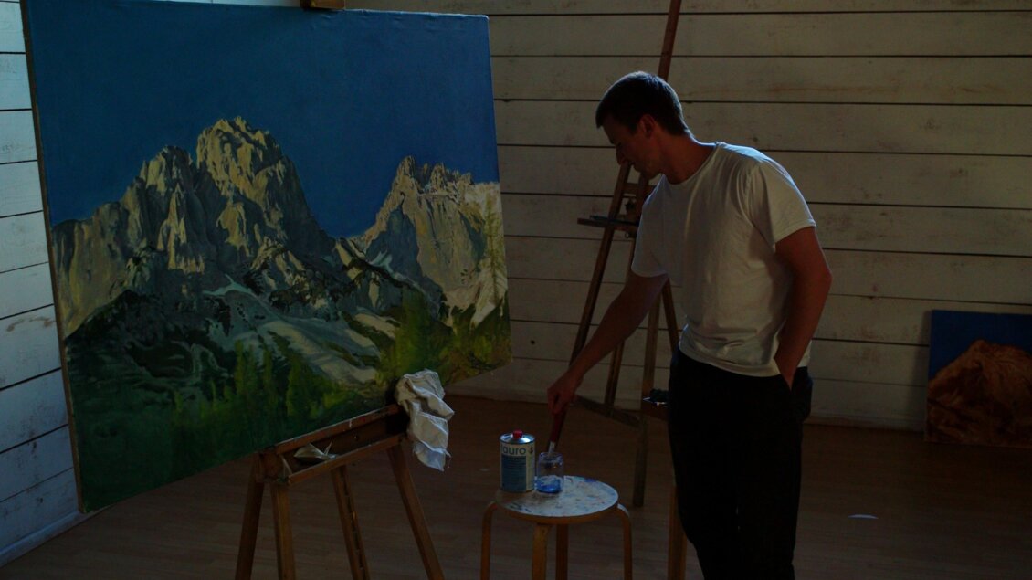 Ein Künstler steht in einem Atelier und arbeitet an seiner Malerei, die Berge zeigt. Auf einem Tisch sind Malutensilien und eine Wasserflasche zu sehen. | © Jakob Brandstätter