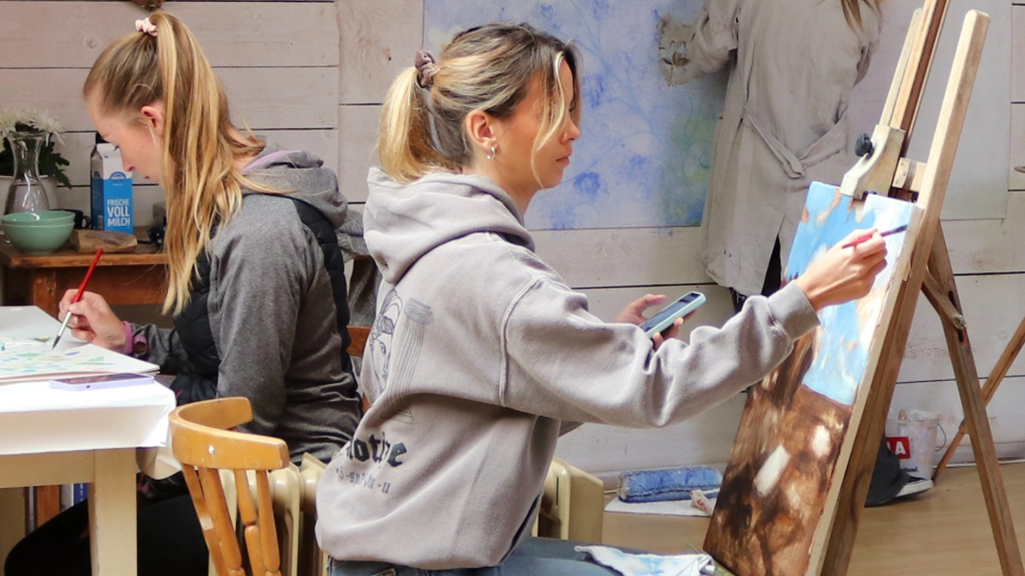 Zwei Frauen malen in einem hellen Atelier. Eine konzentriert sich auf ihre Leinwand, während die andere im Hintergrund eine Wand bemalt. | © Jakob Brandstätter