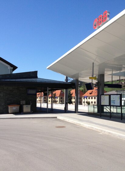 A modern train station with clean lines and an adjacent historic building. The sky is blue and the square is well-lit.