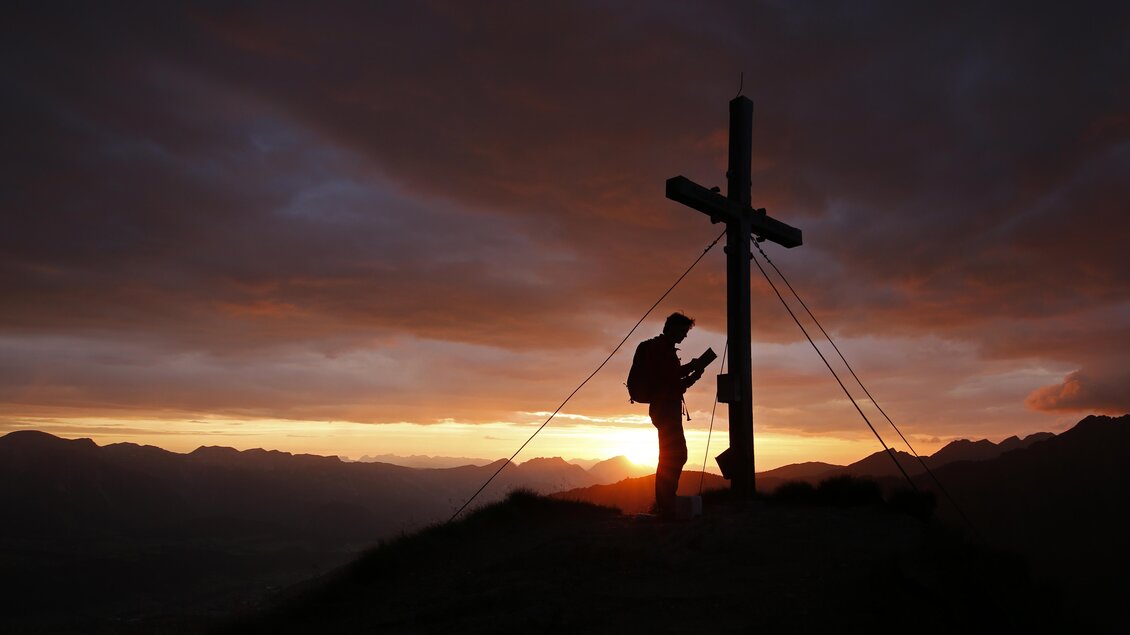 Eine Silhouette eines Mannes, der an einem Gipfelkreuz steht, während die Sonne untergeht. Die Wolken am Himmel sind in warmen Farben beleuchtet. | © Herbert Raffalt