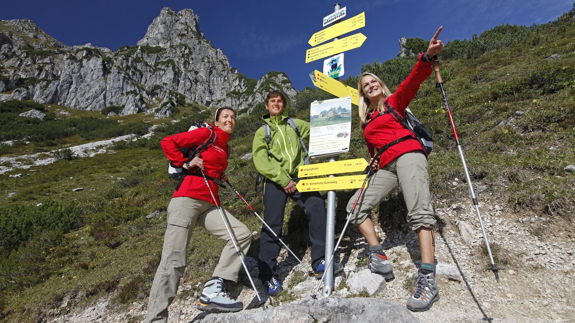 Drei Wanderer stehen an einem Wegweiser in den Bergen. Sie tragen Wanderausrüstung und scheinen sich über die Route zu freuen. | © Raffalt