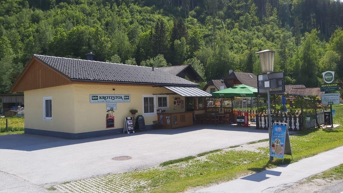 Ein kleines, gelbes Gebäude mit einem Holzschild und einer Terrasse im Freien. Es befindet sich in einer grünen Naturlandschaft mit Bäumen im Hintergrund. | © Sabine Schweinberger
