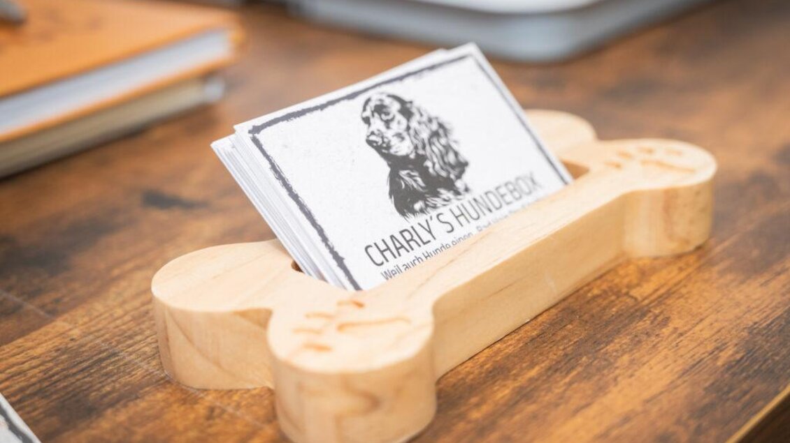 Ein kreativer Ständer in Form eines Hundeknochens hält Visitenkarten mit dem Namen "Charly's Hundebox". Der Hintergrund zeigt eine Holzoberfläche, die eine warme Atmosphäre schafft. | © Tanja Noe