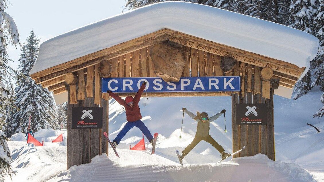 Zwei Personen springen fröhlich unter einem Schild mit der Aufschrift "CROSSPARK". Im Hintergrund ist eine verschneite Winterlandschaft zu sehen. | © lorenzmasser.com