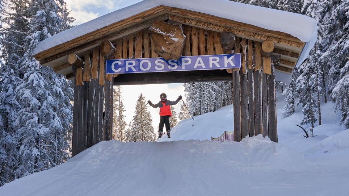 Ein Wintersportort mit einem hölzernen Eingangsschild „CROSSPARK“. Im Vordergrund steht ein Skifahrer in einem roten Outfit auf einer schneebedeckten Piste.