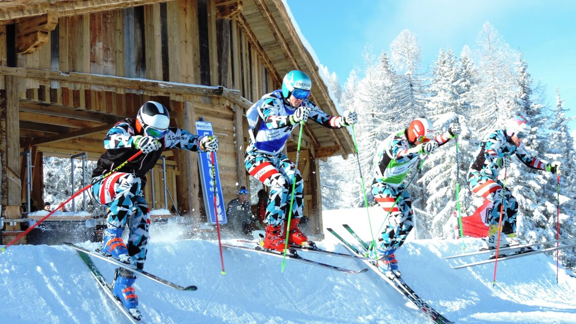 Vier Skifahrer springen im Schnee vor einer Holzhütte. Die Landschaft ist winterlich mit Bäumen und klarem Himmel.