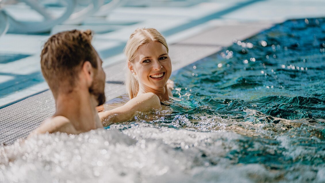Ein Couple entspannt im Wasser eines Pools. Die Frau lächelt fröhlich, während sie mit ihrem Partner plaudert. | © Christine Höflehner Photography