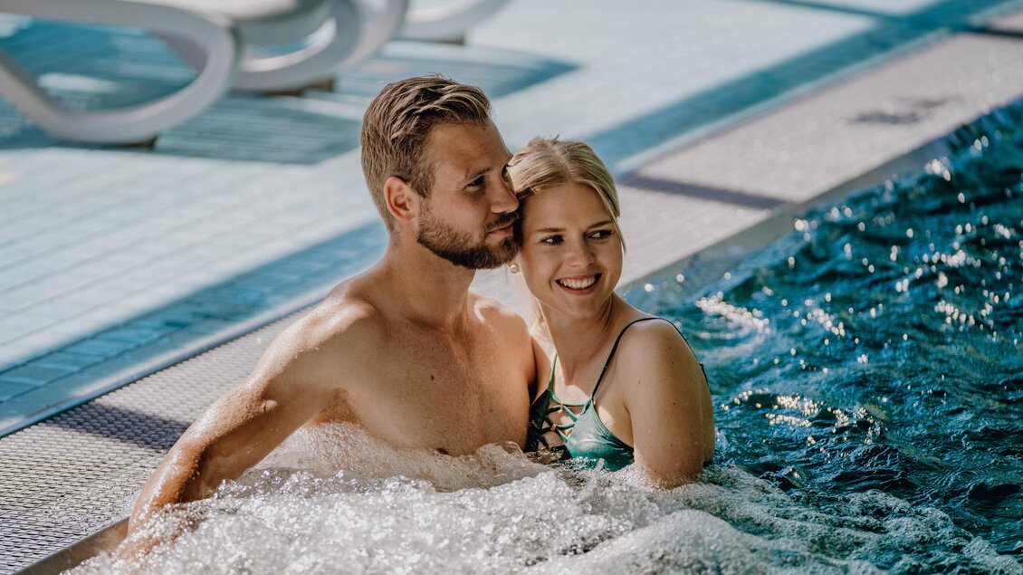 Ein Paar entspannt in einem Pool und genießt die Zeit miteinander. Wasser spritzt sanft um sie herum, während sie lächeln und sich nah sind. | © Christine Höflehner Photography