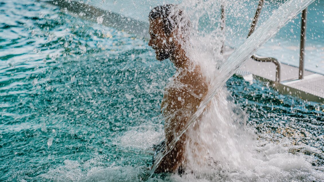 Eine Person steht in einem Pool, während Wasser über sie spritzt. Die Szene vermittelt ein Gefühl von Spaß und Erfrischung. | © Christine Höflehner Photography