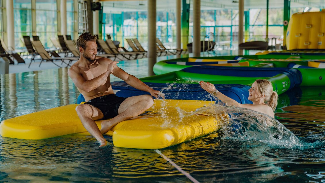 Zwei Personen spielen im Wasser. Einer sitzt auf einer gelben Matratze, während die andere ihn mit Wasser bespritzt. | © Christine Höflehner Photography